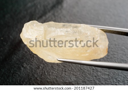 Raw, uncut and rough calcite crystal held by tweezers over black stone.
