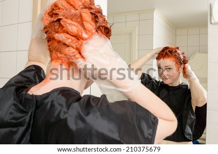 Caucasian woman with short hair dying her hair red in front of mirror in her own bathroom.
