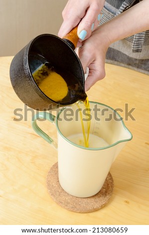 Female hands adding melted butter from cast iron pan into milk in enameled liter measure.