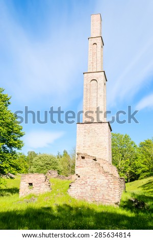 Towering smokestack left standing among ruins after an old factory. Green nature surrounding it. Blue sky with clouds.