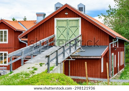 Wooden vehicle and animal ramp leading up to a house on second floor. Red farm building that uses both the upper and lower floor for storing animals and utensils. Ramp rests on part of roof.