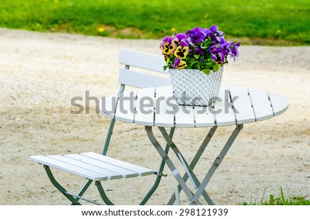 Lovely purple and yellow pansy flowers on white outdoor furniture. Furniture has metal details and wooden surface. One chair and table. Flowerpot has netting on outside. Gravel and grass in background