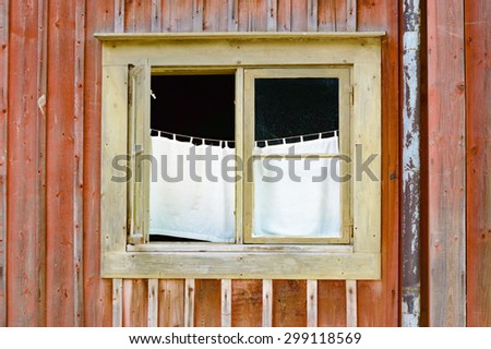 Open window on an old red wooden house. Inside hangs a white very simple curtain from a rope or string. Drainpipe visible beside window. No person in picture. Place has a weathered and run down look.