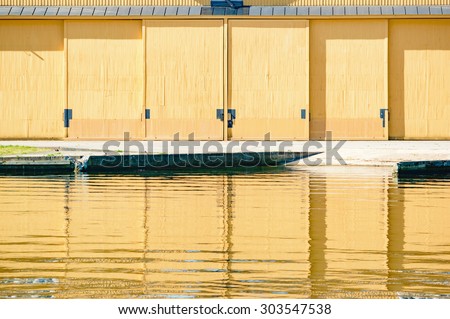 Yellow doors on a hangar are reflected in the calm water in front. Concrete ramp leads up to building from water. Hangar is closed.