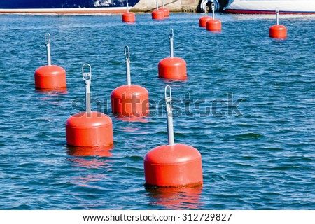 Lots of empty red mooring buoys floating on water in marina. Small part of pier and boats visible at the top of image. Calm water with small waves. No boats at any buoy.
