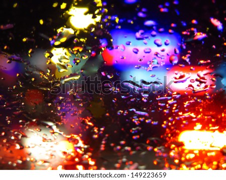 A background of a glass covered with raindrops with a backdrop of vehicle lights at night.