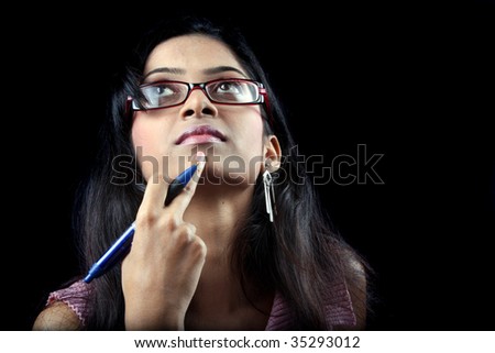 An Indian teenager with a pen trying to remember the answer of a question during her exam, on black studio background.