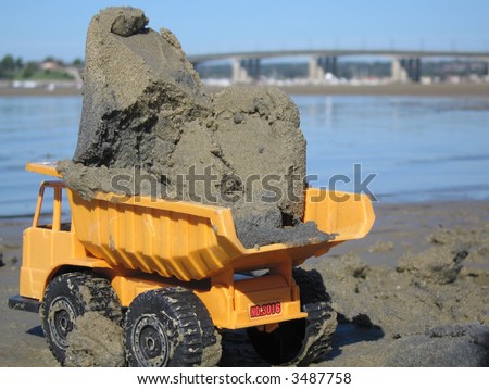 Yellow toy dump truck in the sand carrying a load with view of beach and bridge
