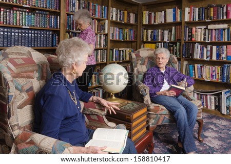Two elderly women talk in a comfortable private library while another women browses in the background
