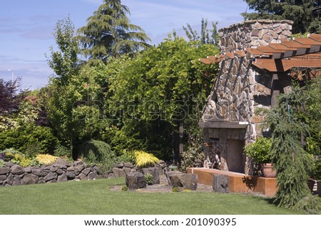 A massive fireplace with sitting stones in front of it in a beautifully landscaped garden with a dry stone wall in the background at the edge of tall arbor walkway.