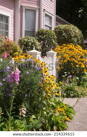 Bright summer flowers almost hide an old fashioned gate to a pink Victorian home.