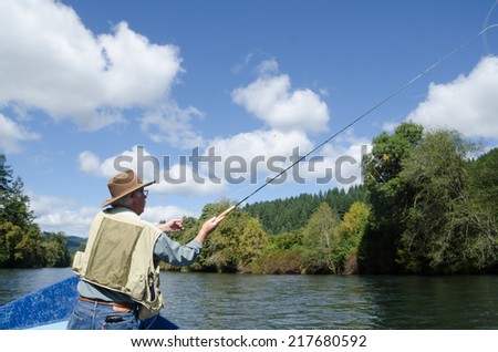 An elderly fisherman in a small boat casts his line under a bright blue summer sky dotted with fluffy white clouds.