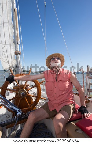 A senior man at the helm of his classic sailboat judges the wind in his sails under a bright blue sky.
