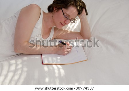 A thirty-something woman lies on a sun-dapple white bedspread writing in a notebook/ Extra space on the bottom and right leave room for designer copy if desired.