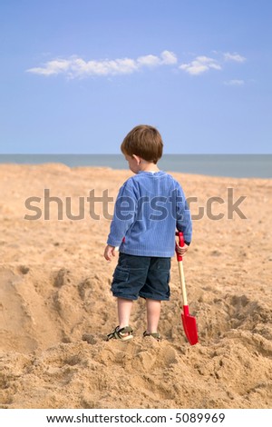Toddler on the beach admiring a huge hole he has just dug in the sand.