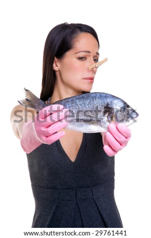 A woman holding a fish at arms length whilst wearing pink rubber gloves and a clothes peg on her nose. Isolated on white background, focus on the fish.