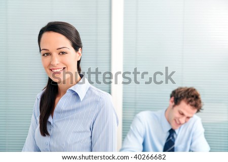 Businesswoman in an office smiling to camera, male colleague in the background out of focus.