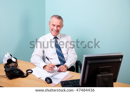 Mature businessman sat at an office desk working on some paperwork smiling to camera. The graphs and text on the documents were made and printed by me.