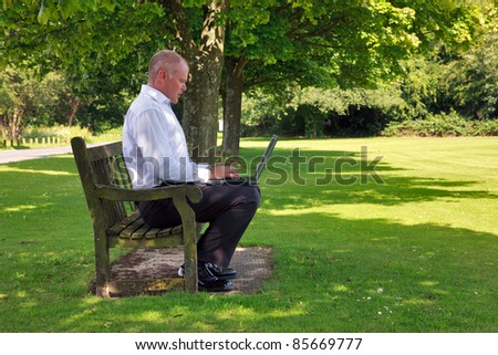 Photo of a businessman in smart casual clothing sat on a park bench working on his laptop computer.