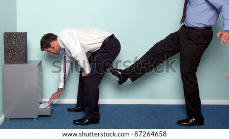 Photo of a businessman bending over to get something out of a drawer as a colleague kicks him up the backside.