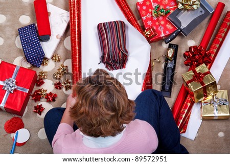 Overhead photo of a woman sat on a rug at home wrapping her Christmas presents surrounded by gifts, paper, ribbon and bows.