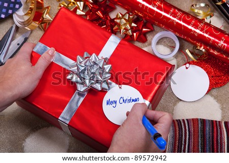 Photo of a woman writing Merry Christmas on a gift tag on a red present with silver ribbon and bow. Of course you could easily add your your message.