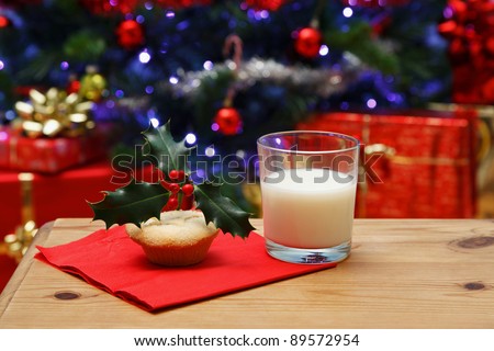 Photo of a glass of milk and a mince pie with holly on top on a table, left out on Christmas eve for Santa, Christmas tree in the background with fairy lights and decorations.