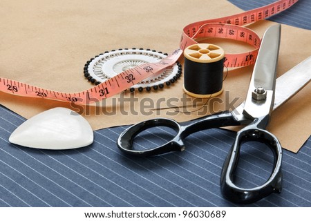 Still life photo of bespoke suit making equipment including tape, chalk, thread, scissors and pins on a pinstripe woolen piece of fabric.