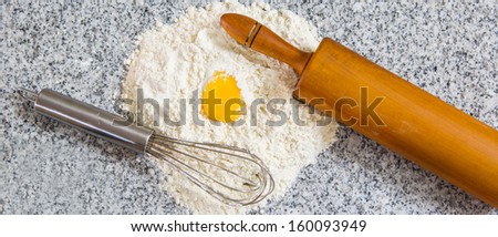 Pastry making preparation with egg, yolk, flour,  egg beater and rolling pin on a granite counter surface.