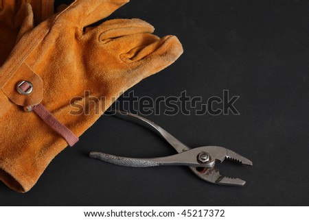 Brown suede leather work gloves and chrome plated slip joint pliers on a black background