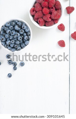 bilberry and raspberry fruits at white ceramic cup, on bright wood table