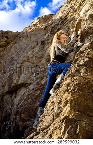 Attractive slim woman in blue jeans blindfolded. Is climbing a big cliff. Leopard print clothes.