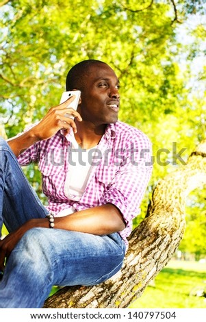 Young smiling african american sitting on a tree in park and calling with mobile phone