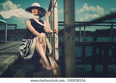 Beautiful woman wearing hat and white scarf sitting on old wooden pier