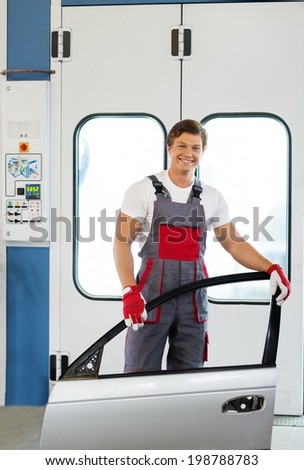 Cheerful serviceman with car door near paint booth in a car body workshop