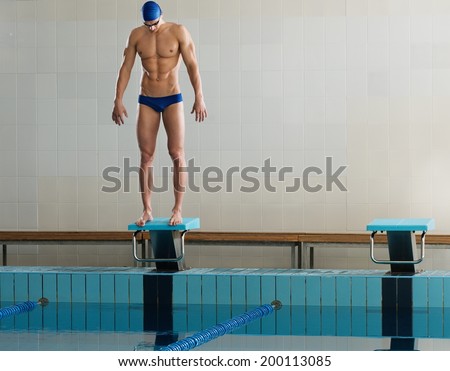 Young muscular swimmer standing on starting block in a swimming pool