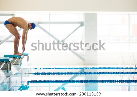 Young muscular swimmer in low position on starting block in a swimming pool