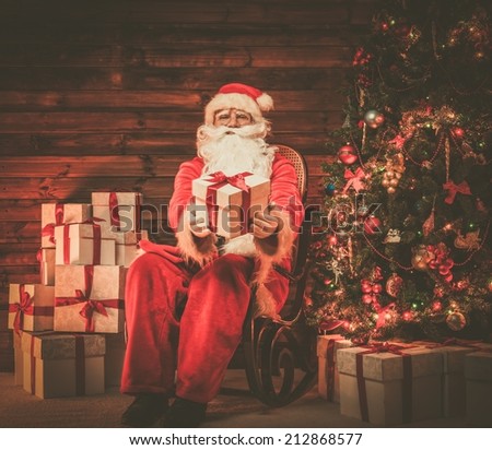 Santa Claus sitting on rocking chair in wooden home interior presenting gift box