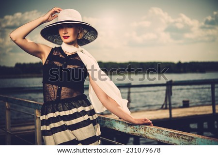 Beautiful woman wearing hat and white scarf sitting on old wooden pier