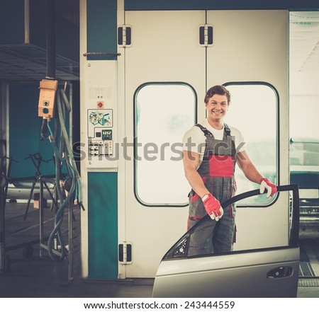 Cheerful serviceman with car door near paint booth in a car body workshop