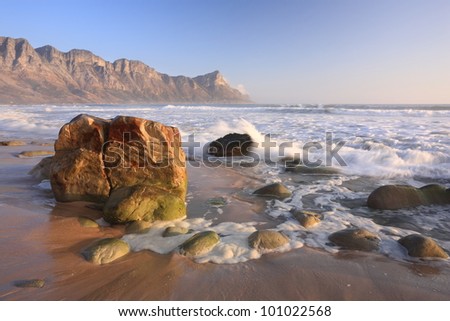 Rocky beach with coastal mountains, blue sky and sand near Cape town, South Africa, in soft evening light.