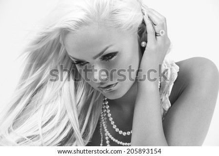 Gorgeous blond woman with a dreamy expression wearing expensive pearl jewellery looking pensively down at the floor with her long hair blowing in the breeze and a wistful expression, greyscale image