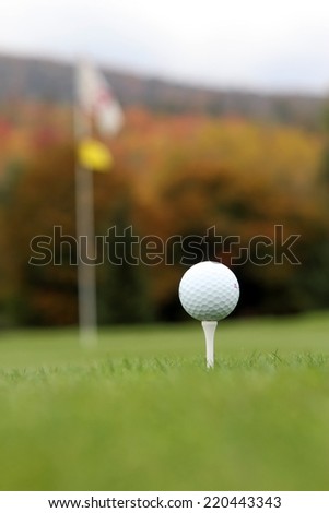 Golf ball on tee with flag stick and autumn foliage in background.