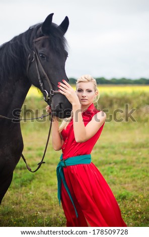 Young cute woman in saturate long dress with black strong and muscular horse in field