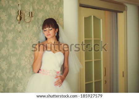 Gorgeous bride in wedding dress in luxury interior with diamond jewelry posing at home and waiting for groom. Romantic rich happy girl in bridal dress