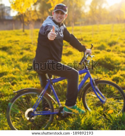 Young man show ok sign by hands and cycling on a rural road through green spring meadow during sunset