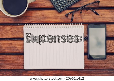 Notebook with text Expenses inside  on table with coffee, mobile phone and glasses.