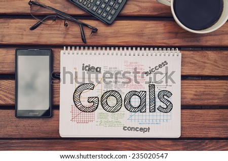 Notebook with text inside Goals on table with coffee, mobile phone and glasses.