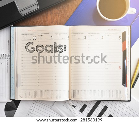 Notebook with text inside Goals on table with coffee, laptop and some sheet of papers with charts and diagrams