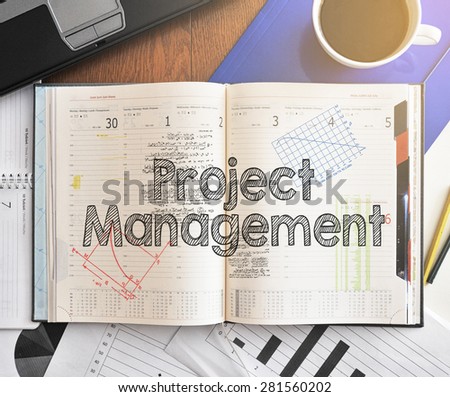 Notebook with text inside Project Management on table with coffee, laptop and some sheet of papers with charts and diagrams